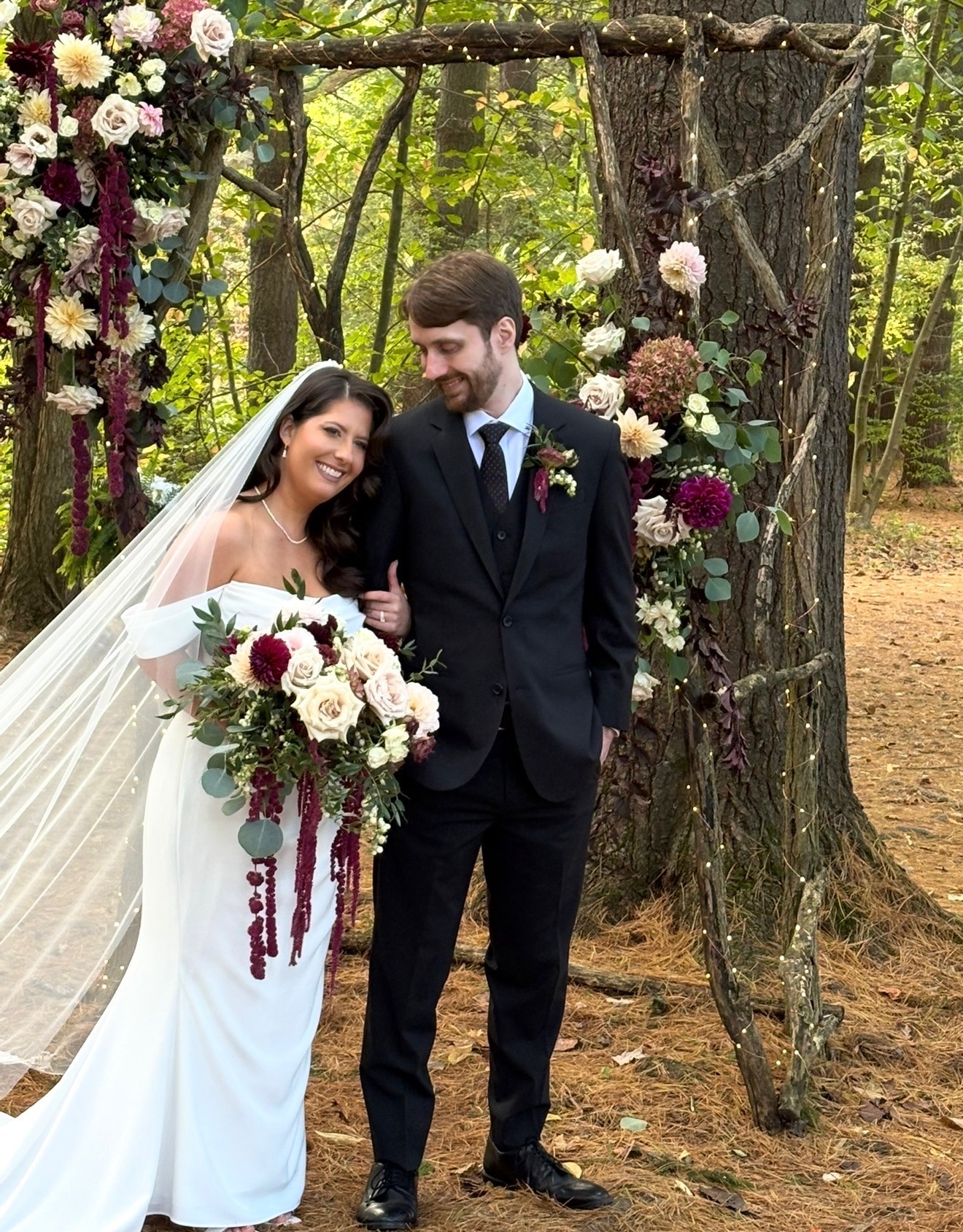 Bride and groom smiling under a floral and greenery arch, forest ceremony